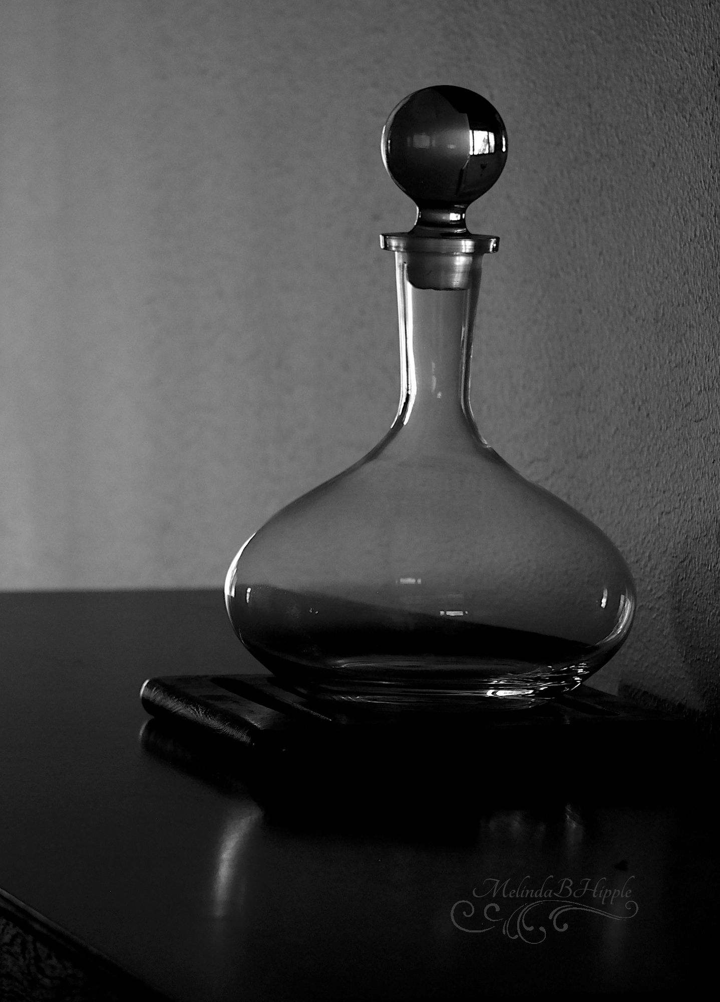 Black and white photo of a clear glass decanter sitting on a square flat base on a tabletop. Window light from off camera is highlighting its edges. The simple design is broad at the base, tapering to a narrow long neck. The clear stopped is a simple round ball.
